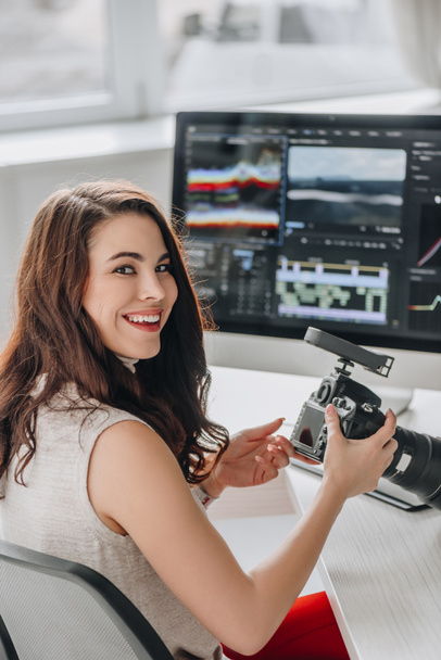 Счастливый редактор, держащий цифровую камеру рядом со столом с компьютерными мониторами
  - Фото, изображение