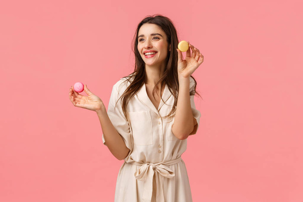 Hübsche junge Unternehmerin, die sich selbstständig macht, Desserts backt, Freunde vorschlägt, Macarons in der Hand hält und fröhlich lächelt, amüsiert nach links schaut, vor pinkem Hintergrund erfreut steht - Foto, Bild