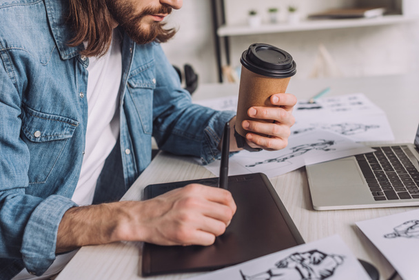 cropped view of bearded animator holding paper cup near sketches  - Photo, Image