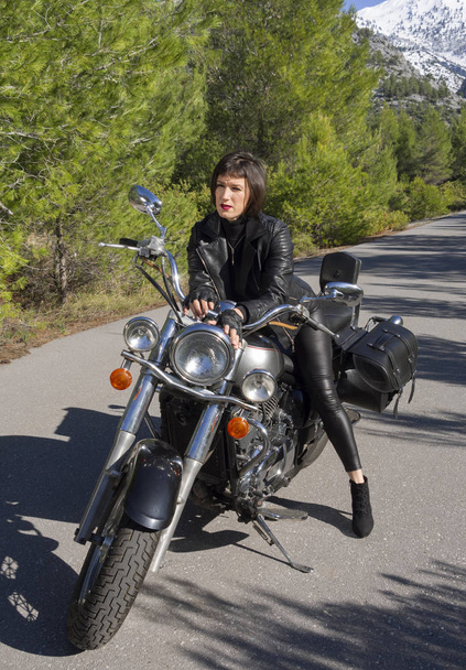 Uma mulher em uma jaqueta de couro preto motociclista em uma motocicleta helicóptero na Grécia em uma estrada na floresta nas montanhas
 - Foto, Imagem