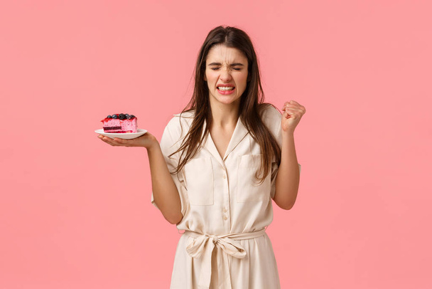 A lány hányni fog a torta gusztustalan ízétől. Csalódott fiatal nő megpróbál enni desszert és grimaszolás, guggoló szörnyű szag, gazdaság desszert és álló rózsaszín háttér - Fotó, kép