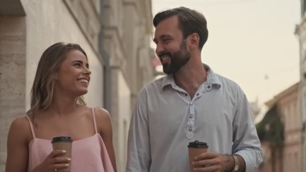 The laughing happy young lovely couple are walking by street outdoors holding paper cups of coffee or tea - Imágenes, Vídeo