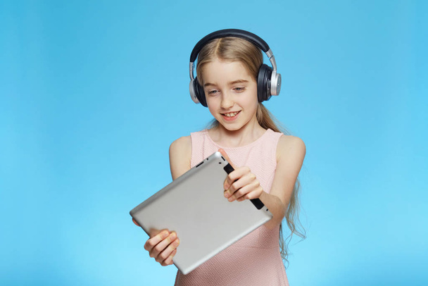 Beautiful little cheerful girl is playing a game on a tablet. Girl wears headphones. Portrait on a blue background. - Photo, Image