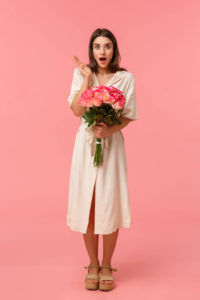 Full-length vertical portrait pretty surprised girl receive unexpected gift, got delivery holding flowers, gasping open mouth amazed and looking speechless with stunned expression, pink background - Fotografie, Obrázek