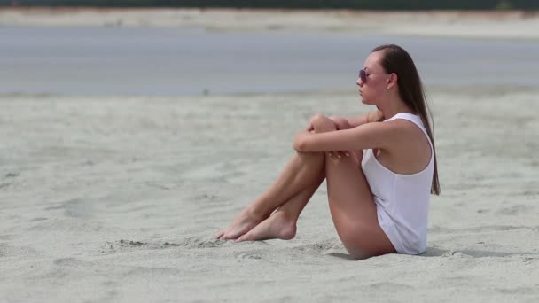 Languid kobieta siedzi Cross-legged na piasku dotykając skóry - Materiał filmowy, wideo