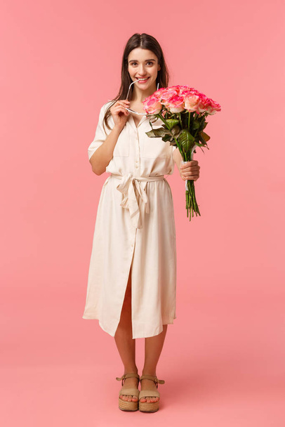 Full-length κάθετο πορτρέτο αισθησιακό, ρομαντικό και coquettish νεαρή γυναίκα βλέποντας κάτι ενδιαφέρον, ψάχνει με πειρασμό ή επιθυμία, κρατώντας λουλούδια μπουκέτο, έλαβε τριαντάφυλλα, ροζ φόντο - Φωτογραφία, εικόνα