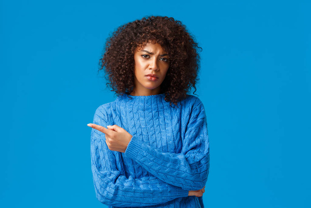 Explain to me, what is this. Displeased gloomy and unsatisfied african-american girlfriend with afro haircut, pointing finger left, frowning skeptical and disapproval, blue background - Photo, Image