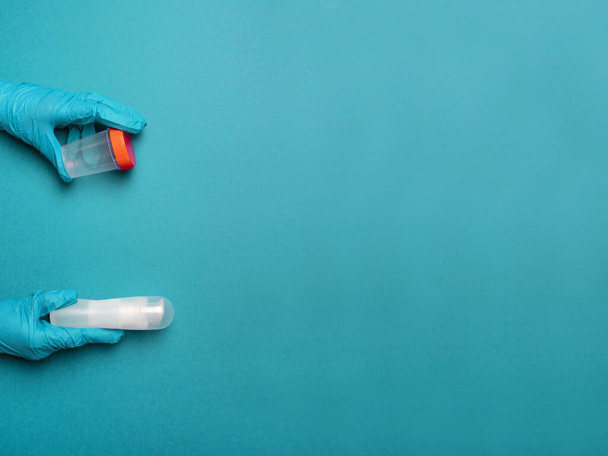 Ärztehände in Latex-Gummihandschuhen, die einen Plastikbehälter und ein Schmiermittel mit Flüssigkeiten auf blauem Hintergrund mit Platz für Text halten - Foto, Bild