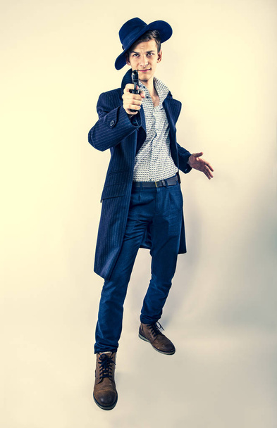 Mysterious undercover young man-gangster aiming a gun at the camera and wearing a dark hat and coat isolated on light background - Foto, immagini