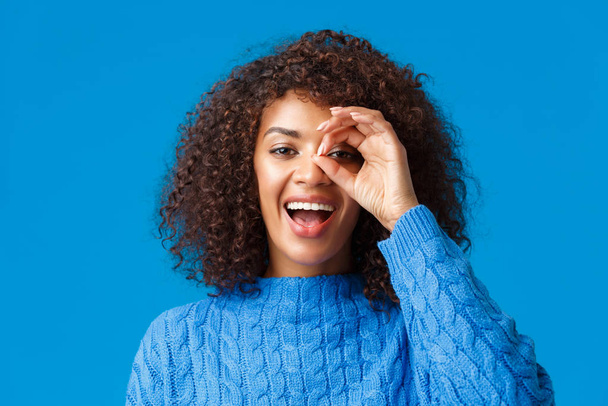 Looking into distance. Close-up cheerful attractive african-american woman searching something, found great holidays discount, look through okay sign and smiling pleased, blue background - Foto, imagen