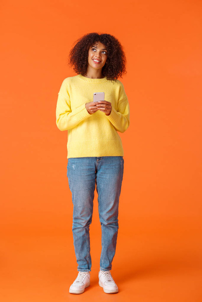 full-length vertical tiro sonhador bonito afro-americano mulher pensando-se o que escrever, segurando smartphone pensando olhando para cima e sorrindo coisas de imagem, de pé fundo laranja alegre
 - Foto, Imagem
