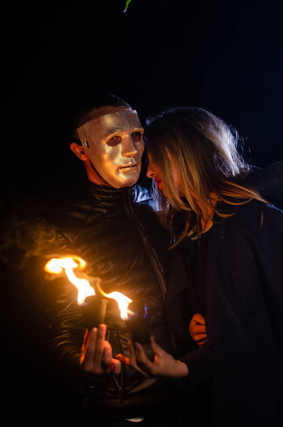 tajemnicza młoda dziewczyna w czarnym płaszczu i mężczyzna w masce, sabat wiedźmy w lesie w nocy przy ognisku - Zdjęcie, obraz
