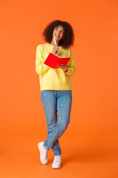 Full-length vertical shot dreamy and romantic cute girl making schedule, taking notes or to-do list, imaging something as writing in red cute notebook, touch chin with pen looking up thoughtful - Foto, imagen