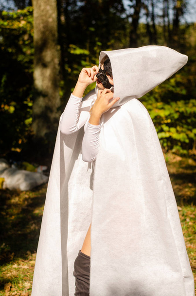 girl wearing a white raincoat and black mask, carnival forest. - Foto, afbeelding