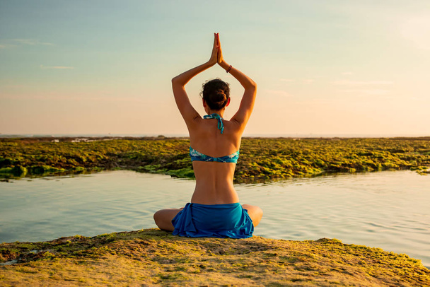 junge Frau, meditiert, praktiziert Yoga und Pranayama am Strand. Yoga bei Sonnenuntergang. Hände, die sich in namaste mudra erheben. Blick von hinten. Strand von Melasti, Bali - Foto, Bild