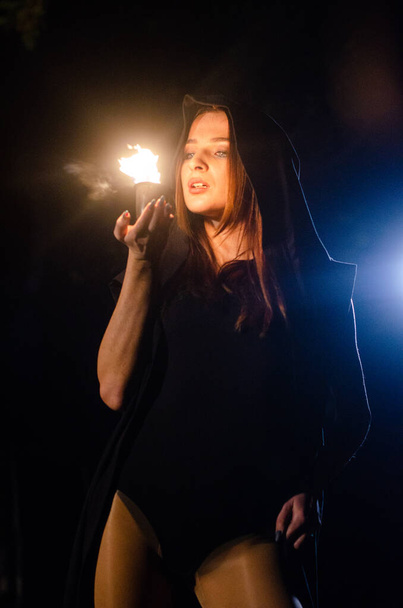 jovem misteriosa vestindo um manto preto, sábado de bruxa na floresta à noite perto da fogueira - Foto, Imagem