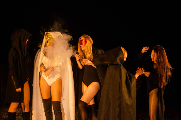 μυστηριώδη νεαρά κορίτσια που φοράνε ασπρόμαυρες μανδύες, το Σάββατο των μαγισσών στο δάσος τη νύχτα δίπλα στη φωτιά. - Φωτογραφία, εικόνα
