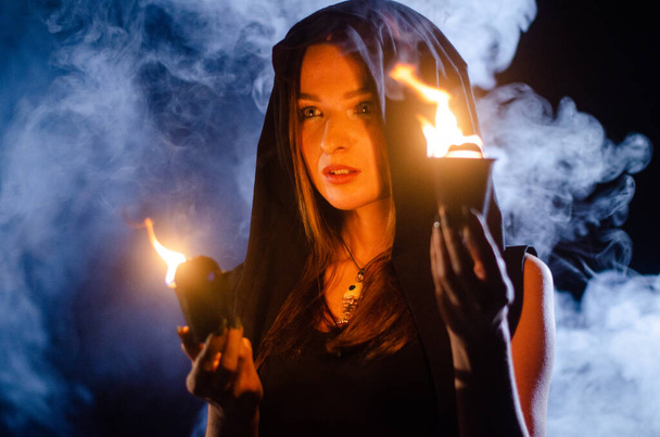 jovem misteriosa vestindo manto preto, sábado de bruxa na floresta à noite perto da fogueira - Foto, Imagem