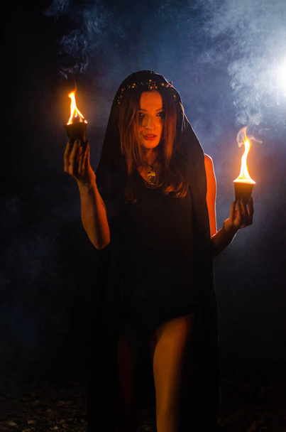 Siyah pelerin giyen gizemli genç kız, Şenlik ateşinin olduğu gece ormanda cadı ayini. - Fotoğraf, Görsel