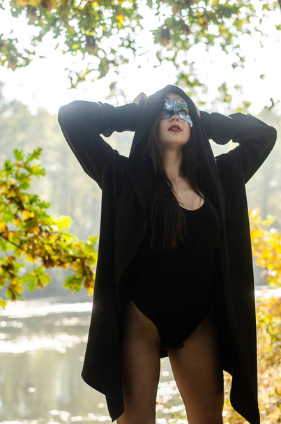 beautiful young woman in black carnival costume and mask posing on the woods - Photo, Image