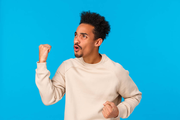 Lottery, sport bet, people concept. Tensed and excited young handsome african-american guy watching game on tv in pub, looking left screen, fist pump rooting for team, triumphing, blue background - Foto, Imagem