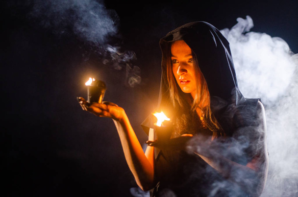 misteriosa jovencita vestida con capa negra, sábado de brujas en el bosque por la noche junto a la hoguera - Foto, Imagen