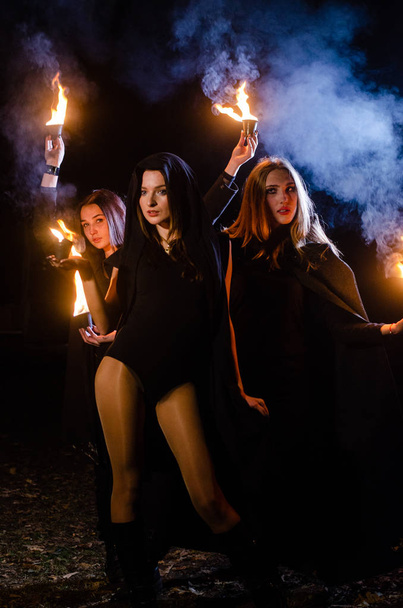tajemnicze młode dziewczyny w czarnych płaszczach, sabat czarownic w lesie w nocy przy ognisku - Zdjęcie, obraz