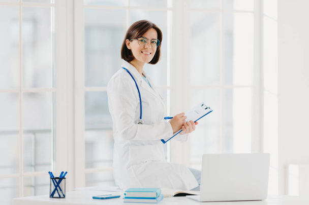 Ärztin sitzt auf Tisch, notiert Informationen in Klemmbrett, posiert am Arbeitsplatz im Krankenhausbüro, arbeitet mit Laptop, lächelt freundlich. Gesundheit, Technologie, Medizin, Internetkonzept - Foto, Bild