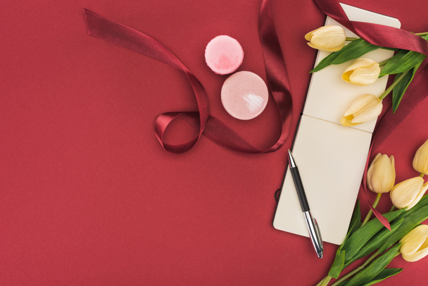 top view of tulips with silk ribbon, macarons and empty notebook with pen isolated on red - Fotografie, Obrázek