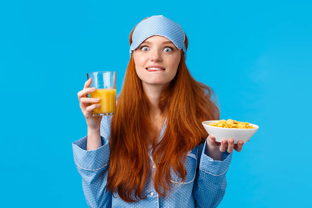 Восхитительно. Женщина ест любимую еду по утрам. Милая рыжеволосая студентка в маске для сна и ночной одежде, облизывающая губы от соблазна и удовлетворения, поедающая вкусные хлопья и апельсиновый сок
 - Фото, изображение