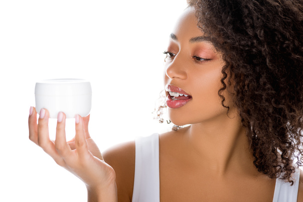 junge lächelnde afrikanisch-amerikanische Frau hält Plastikbehälter mit Gesichtscreme, isoliert auf weiß - Foto, Bild