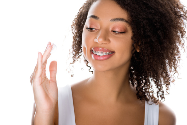 chica afroamericana sonriente con frenos dentales aplicando crema facial, aislado en blanco
 - Foto, Imagen