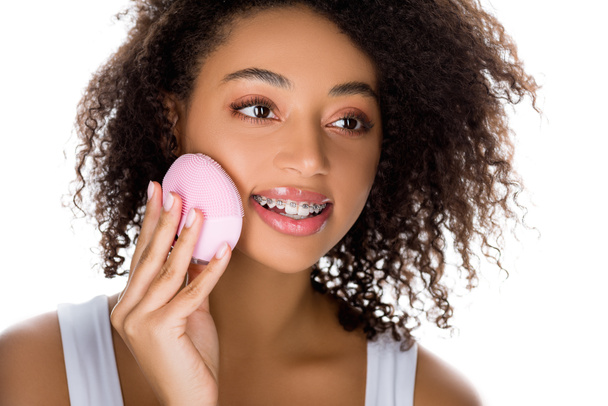 attraktive glücklich afrikanisch-amerikanische Mädchen mit Zahnspangen mit Silikon Reinigung Gesichtsbürste, isoliert auf weiß - Foto, Bild