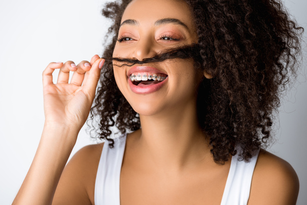 Смеющаяся африканская американка с зубными брекетами, делающая фальшивые усы из волос, изолированная от серого
 - Фото, изображение