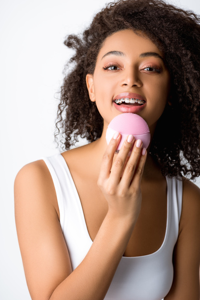 happy african american woman with braces using silicone cleansing facial brush, isolated on grey - Photo, Image