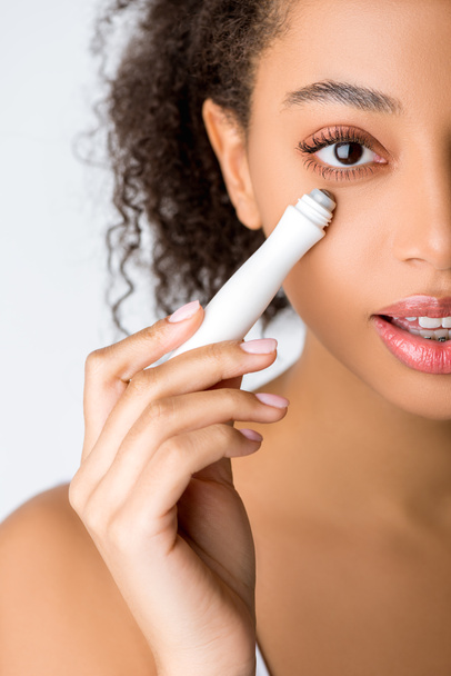 curly african american girl using moisturizing eye roller, isolated on grey - Photo, Image