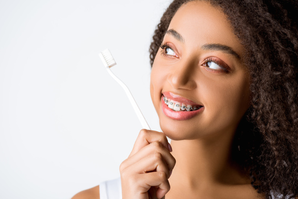beautiful curly african american woman with dental braces looking at toothbrush, isolated on grey - Foto, Bild