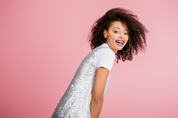 smiling african american girl with dental braces, with silver glitter eyeshadows and purple lips dancing in paillettes dress, isolated on pink - Foto, Imagem