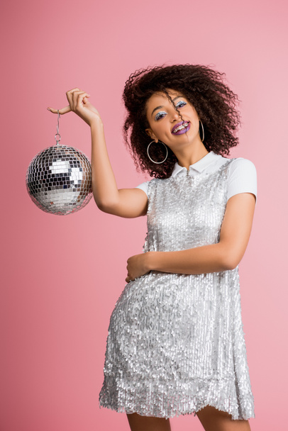 cheerful african american girl in paillettes dress holding disco ball, isolated on pink  - Fotó, kép