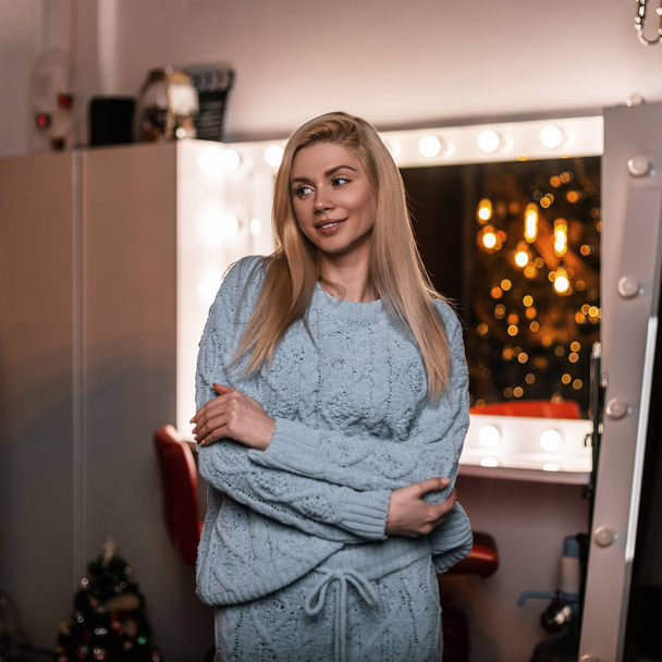 Cute young beautiful blond woman with a lovely smile posing in a blue fashionable knitted suit in a room with a New Year tree. Attractive trendy happy girl in the studio near a white makeup mirror. - Photo, Image