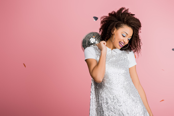 smiling african american girl in paillettes dress holding disco ball, isolated on pink with confetti - Fotoğraf, Görsel