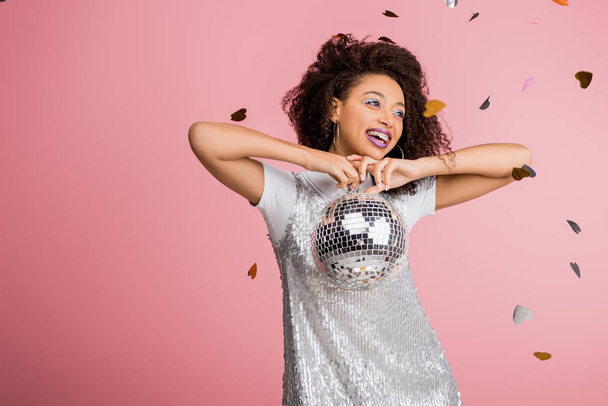happy african american girl in paillettes dress holding disco ball, isolated on pink with confetti - Fotografie, Obrázek