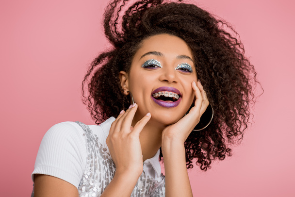 excited african american girl with dental braces, with silver glitter eyeshadows and purple lips wearing paillettes dress, isolated on pink - Photo, Image