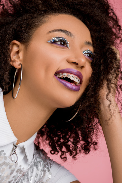 cheerful african american girl with dental braces, with silver glitter eyeshadows and purple lips wearing paillettes dress, isolated on pink - Photo, Image