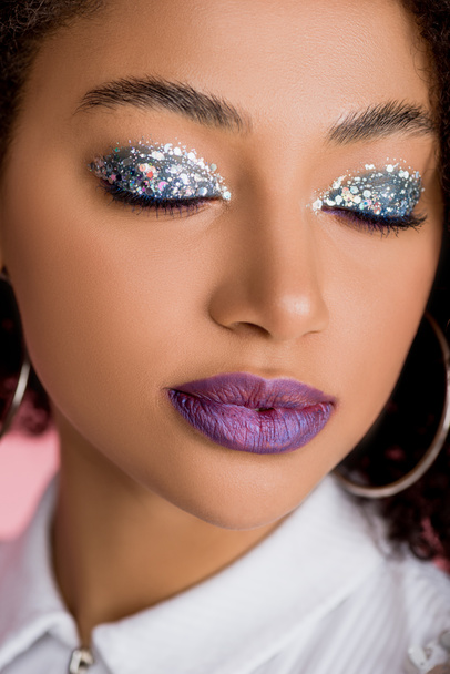Afrikaans Amerikaans meisje met zilveren glitter oogschaduwen op gesloten ogen en paarse lippen, geïsoleerd op roze - Foto, afbeelding