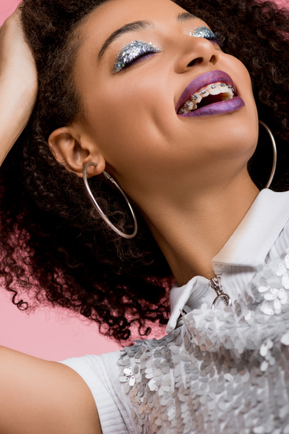 happy african american girl with dental braces, with silver glitter eyeshadows and purple lips wearing paillettes dress, isolated on pink - 写真・画像