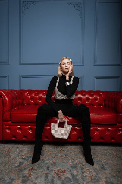Стильная красивая молодая женщина в стильной черной одежде с белой элегантной жемчужной сумкой в винтажном перламутровом топе сидит на кожаном красном диване в помещении.
 - Фото, изображение