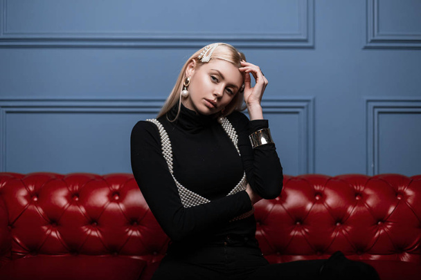 Modern glamorous young woman posing in a black shirt in a fashionable white pearl top with elegant pearl hair clips indoors near a blue wall. Stylish girl fashion model sits on a red sofa in the room. - Foto, imagen