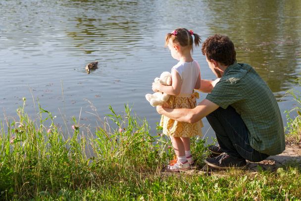 Baba ve kızı göle bakıyor. Kızın elinde oyuncak ayı var. Vahşi bir ördek suda yüzüyor. Arka plan. - Fotoğraf, Görsel