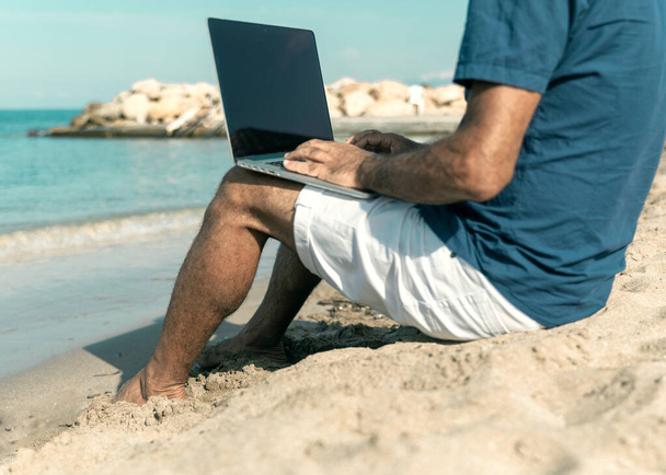 A man sitting on sandy beach and working on computer at warm summer day. Freedom and travel concept - Фото, изображение